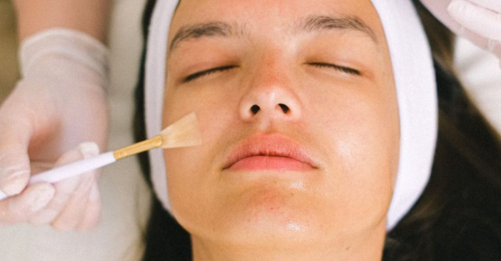 Spa Services - Top view skilled female cosmetologist in uniform and gloves applying essential oil on young woman face skin while working in contemporary spa center